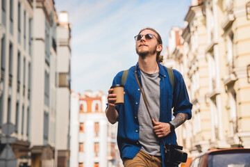 Handsome young man in casual clothing enjoying summer and wandering empty streets while walking outdoors with cup of coffee in hands. Solo trip city exploration
