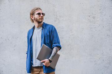 Portrait of a handsome young businessman leaning against a grey wall holding a laptop. Caucasian freelancer student remote worker using computer for remote work, e-learning, coding, it