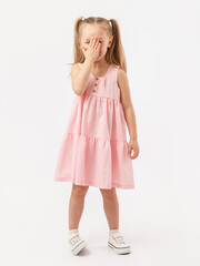 A 5-year-old girl with long hair in a summer pink dress stands on a white background, covering her face with her palm.