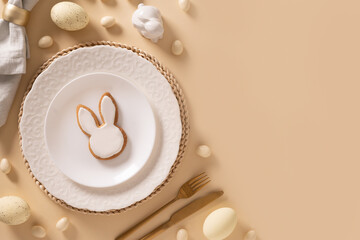 Easter table setting background with white plate, cookies , eggs and bunny on beige background. View from above. Space for greeting text.