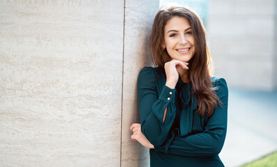 Charming woman in an elegant green dress leaning on a textured wall, smiling casually.