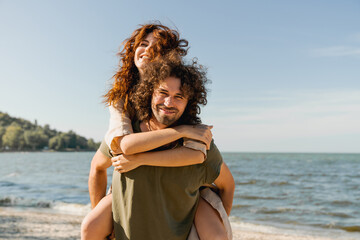 Handsome young man giving piggyback ride to girlfriend on beach while enjoying holidays