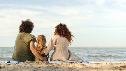 Fototapeta premium Rear view shot of happy family enjoying the weekend, sitting on blanket on a sunny beach