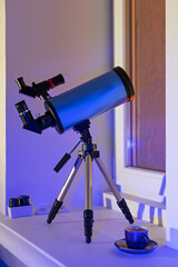 A modern telescope stands on the windowsill and is pointed out the window. Blue catadioptric...