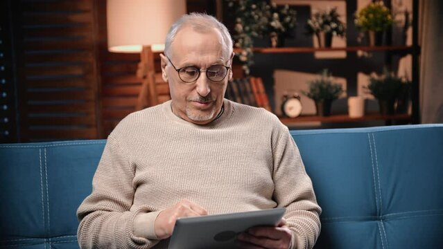 Joyful senior man using tablet for surfing internet at home, swiping and scrolling display, portrait