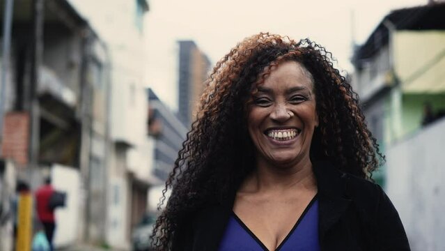 One happy middle aged Brazilian black woman smiling and laughing while walking forward toward camera in urban city street environment, authentic real life laugh and smile
