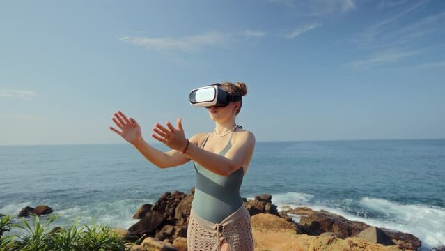 Woman interacts with virtual reality headset by seaside, gestures as if in game or exploring digital world. Engaged in VR adventure, waves crash behind. Slow motion.