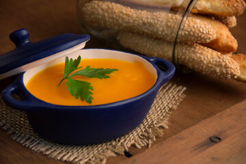 carrot soup with a parsley leaf in a small blue ceramic pot, on top of a jute, in the background crotons in a transparent glass jar, rustic wooden table, horizontal photo, front © Giovanna