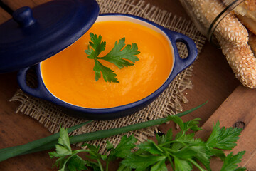 carrot soup with a parsley leaf in a small blue ceramic pot, on top of a jute, in the background crotons in a transparent glass jar, rustic wooden table, horizontal photo, top © Giovanna