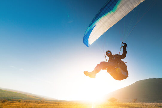 A paraglider takes off from a mountainside with a blue and white canopy and the sun behind. A paraglider is a silhouette. The glider is sharp, with little wing movement.