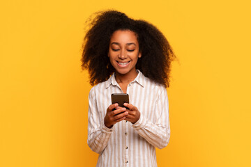 Happy young black woman texting on smartphone against yellow background