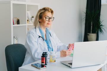 Friendly female middle aged doctor shows jar of pills and pills organizer to laptop in clinic...