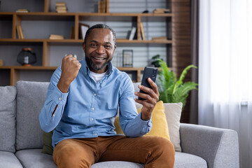 Black skinned man in casual outfit sitting on couch with modern smartphone and making winner gesture in living room. Portrait of mature male rejoicing great news with satisfied facial expression.