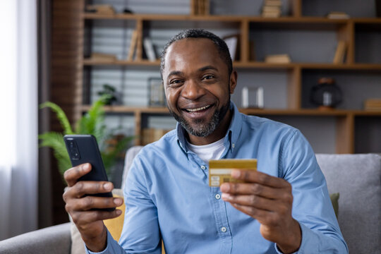 Delighted black male smiling at camera with cell phone and gold credit card in hands on apartment interior background. Bearded man using online banking application for cashless remote payments.