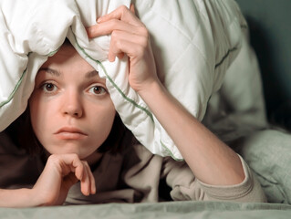 Young girl hugs a pillow and hid under a blanket from all problems, procrastinating, millennials generation. Woman covered with a blanket and is worried about insomnia, poor sleep, getting stress.