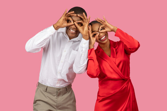 Couple making fun hand gestures over eyes, playful poses, pink background