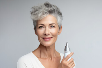 mature woman with silver hair smiles confidently while holding a skincare serum, white background