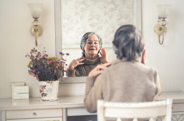 Beautiful old woman sits near the mirror in her cozy light room.