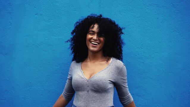 One happy young black Brazilian woman authentic real life laugh and smile standing in outdoor blue backdrop in city street smiling at camera, empowered curly hair African descent person