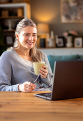 Smiling young woman working at home using laptop while sitting in her living room. - obrazy, fototapety, plakaty