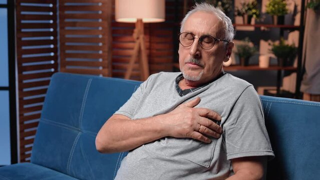 Aged man feeling heartache suddenly and holding left half of chest sitting in evening in home