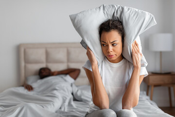 african lady covering ears suffering from husband snoring in bedroom