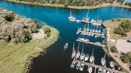 Aerial view of a peaceful marina with various sailboats docked along the pier, surrounded by lush greenery on a bright sunny day - obrazy, fototapety, plakaty