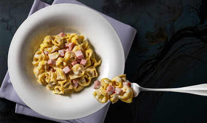 White soup plate and fork with tortellini with cream and ham on black background
