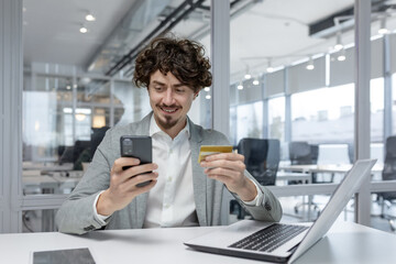 Experienced young adult businessman in a modern office, smiling as he uses a phone. Shows multitasking, focus, and a positive emotional state.