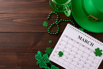 Getting set for St. Patrick's festive fun. Top view photo of calendar, traditional hat, party glasses, green beer, trefoils, beads on wooden background with advert area