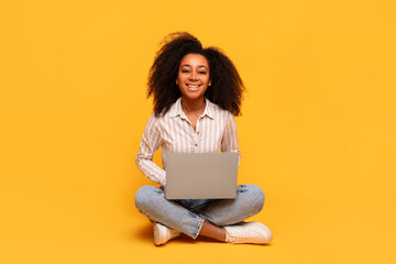 Content black woman with laptop sitting cross-legged on yellow backdrop