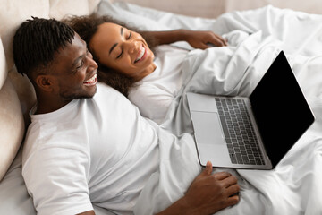Happy young black couple relaxing in bed using laptop indoor - Powered by Adobe