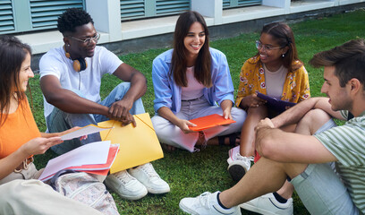 Group of young multiethnic students sitting together outdoors on green lawn at high school. Friends gathering to do schoolwork as a team outside campus university, sitting on the grass.