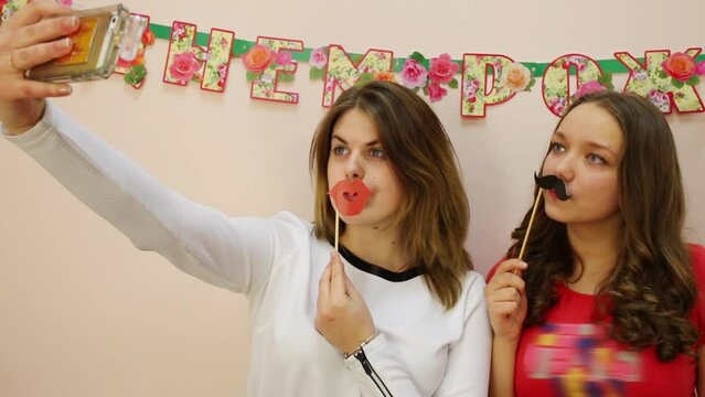 Two girls make selfie with cardboard lips and mustache.