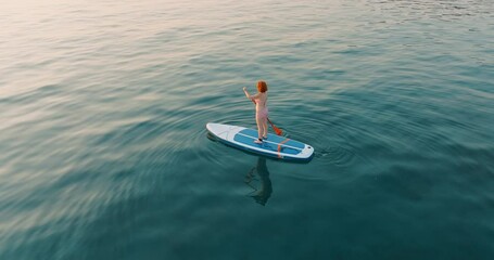 Aerial view young woman floats paddle board on calm blue surface of sea water along coast in Greece at sunset backdrop of hotels in resort town. Summer holiday vacation concept. Lifestyle.Travel - Powered by Adobe
