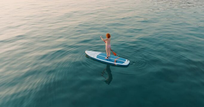 Aerial view young woman floats paddle board on calm blue surface of sea water along coast in Greece at sunset backdrop of hotels in resort town. Summer holiday vacation concept. Lifestyle.Travel