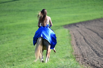 Zwei Sch&ouml;nheiten. M&auml;dchen im blauen Kleid reitet &uuml;ber Blumenwiese