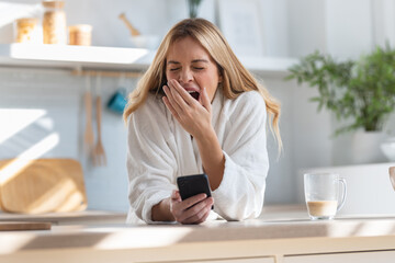 Tired young woman yawning while using her mobile phone in the kitchen at home. - obrazy, fototapety, plakaty
