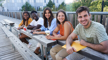 Cheerful group of multiethnic student friends sitting outside of university gathering to do schoolwork together, looking at the camera smiling. 