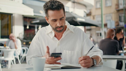 Fototapeta na wymiar Focused freelancer writing notebook at cafe table close up. Guy holding phone