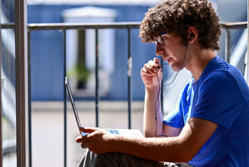 Nice shot of a young Caucasian man working with his laptop talking on earphones. He is sitting on...