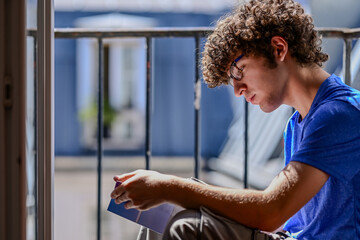 Nice shot of a young Caucasian man reading a book. He is sitting on an attic window, facing the...
