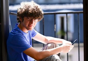 Nice shot of a young Caucasian man with his cell phone. He is sitting on an attic window, he looks into the camera smiling. Natural light illuminates his face and hair. Self-confident and positive.