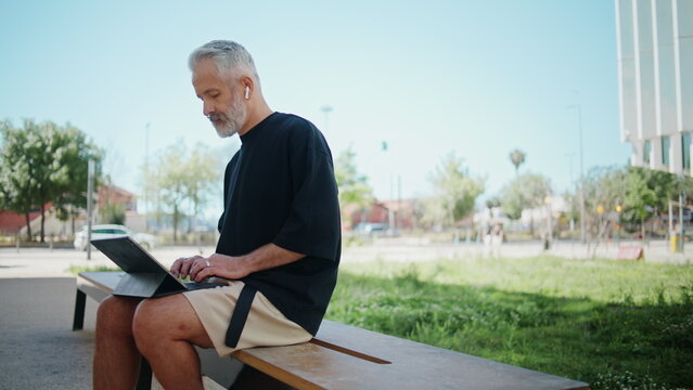 Stylish senior typing computer in urban park. Thoughtful man resting outdoors