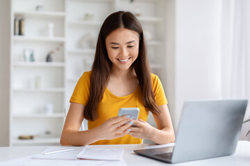Young Asian Woman Using Mobile Phone While Sitting At Desk With Laptop