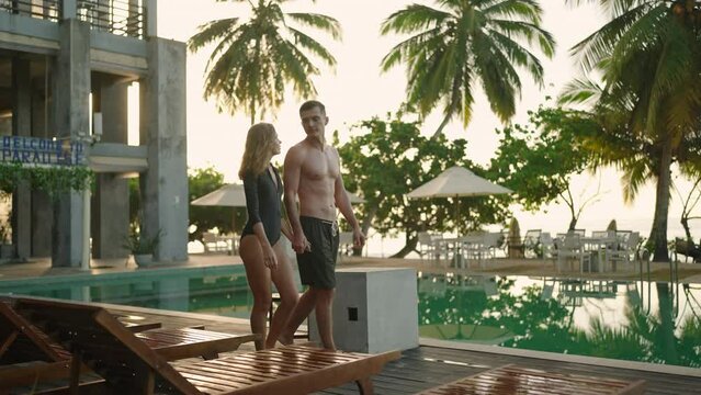Young couple in swimsuits walking along swimming pool of luxury resort hotel on ocean shore. Cheerful girl and guy holding hands near swimming pool. Happy two people relax on poolside together.