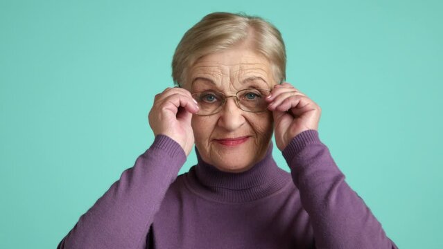 Portrait of cheerful senior lady with blonde short hair, bright lipstick, wearing purple turtleneck, standing over blue background, happy female pensioner. High quality 4k footage