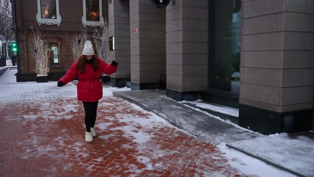 Young woman wearing hat and red trendy winter jacket walking on slippery city sidewalk. Pretty female walking along ice pavement. Danger of season trauma. Shooting in slow motion.