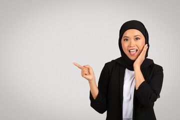 A cheerful young woman in a black hijab and blazer is pointing to the side with one hand while...