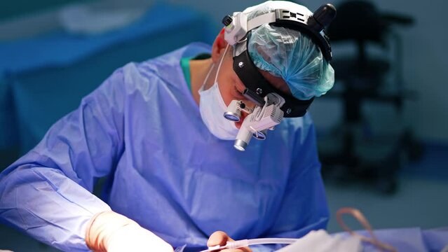 Mid-aged male surgeon working at operation. Professional wearing device glasses uses tool at neurological operation.